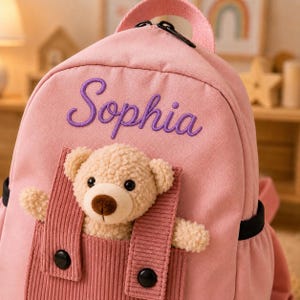 May include: A light pink backpack featuring the name Sophia embroidered in purple script. The front includes a corduroy pocket containing a plush beige teddy bear with black eyes and a brown nose, secured by two black buttons on the straps.