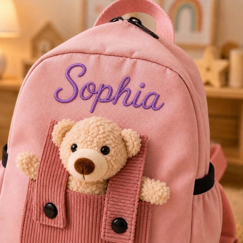May include: A light pink backpack featuring the name Sophia embroidered in purple script. The front includes a corduroy pocket containing a plush beige teddy bear with black eyes and a brown nose, secured by two black buttons on the straps.