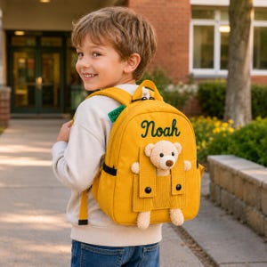 May include: A mustard yellow backpack featuring the name Noah embroidered in dark green script. The bag includes a front pocket with a corduroy flap and a plush beige teddy bear peeking out. A small green tag with the word HAPPY is attached.