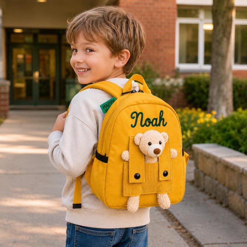 May include: A mustard yellow backpack featuring the name Noah embroidered in dark green script. The bag includes a front pocket with a corduroy flap and a plush beige teddy bear peeking out. A small green tag with the word HAPPY is attached.