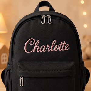 May include: A black backpack featuring the name Charlotte embroidered in light pink cursive script on the front. The bag includes a main zippered compartment with two silver metal pulls and a smaller front zippered pocket with a single silver pull.