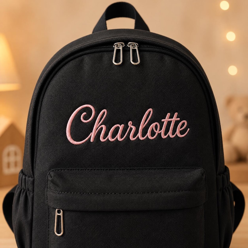 May include: A black backpack featuring the name Charlotte embroidered in light pink cursive script on the front. The bag includes a main zippered compartment with two silver metal pulls and a smaller front zippered pocket with a single silver pull.