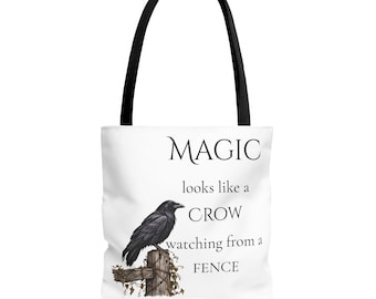 Puede incluir: Un bolso tote blanco con asas negras que presenta un cuervo negro posado sobre un poste de valla de madera con enredaderas. El texto dice: MAGIC looks like a CROW watching from a FENCE. El diseño está centrado en la parte frontal del bolso.