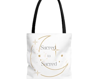 Puede incluir: Un bolso tote blanco con asas negras que presenta un gráfico de luna creciente dorada, iconos de estrellas pequeñas y el texto "Sacred to Sacred" impreso en una fuente serif oscura. El diseño está centrado en la parte frontal del bolso.