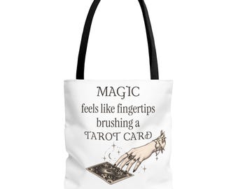 Puede incluir: Un bolso tote blanco con asas negras que presenta el texto MAGIC feels like fingertips brushing a TAROT CARD. Debajo del texto hay una ilustración de una mano con anillos que toca una carta del tarot rodeada de estrellas y lunas pequeñas.