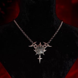 May include: A silver-toned metal pendant necklace featuring a sharp, multi-pointed star design with a central circular element. The pendant is suspended from a delicate silver-toned chain, showcasing intricate, pointed edges and a metallic finish.