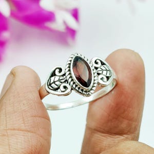 Könnte beinhalten: Ein silberner Ring mit einem dunkelroten, ovalen Edelstein in der Mitte, umrahmt von einem dekorativen Rand. Der Ring hat blattartige Designs auf beiden Seiten des Edelsteins. Der Ring wird von einer Hand gehalten.