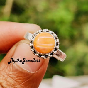 May include: A silver ring featuring a round, orange gemstone with a beaded border. The ring is held by a hand, with the text "Dipika Jewellers" visible on the finger. The gemstone has a striped pattern.