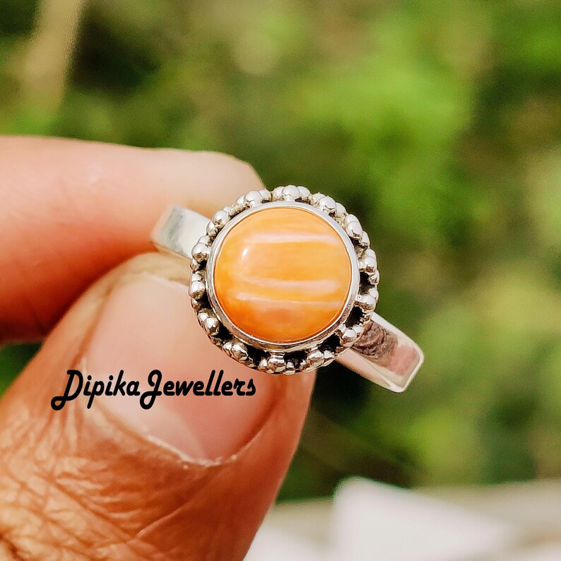 May include: A silver ring featuring a round, orange gemstone with a beaded border. The ring is held by a hand, with the text "Dipika Jewellers" visible on the finger. The gemstone has a striped pattern.