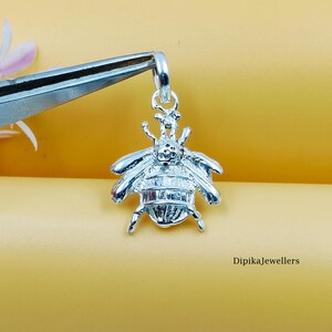 Puede incluir: Un colgante de abeja plateada con alas y cuerpo detallados, sujeto con unas pinzas. El colgante tiene un lazo para una cadena. El fondo es de color amarillo liso. El texto "DipikaJewellers" es visible.