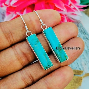 Puede incluir: Un par de pendientes rectangulares turquesa con marcos y ganchos de metal plateado. Los pendientes se sostienen en la mano de una persona. El texto "DipikaJewellers" es visible en la mano.