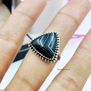 May include: A triangular ring featuring a black and white banded agate stone set in a silver bezel with a beaded edge. The stone displays natural concentric patterns. The text DipikaJewellers is visible on the lower right side of the image.