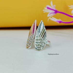 May include: A silver-colored ring with a wing design. The ring features two stylized wings, each with detailed feather patterns. The wings are positioned to create a unique, open design. The ring is set against a soft, neutral background.