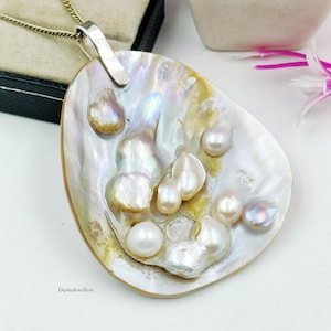 May include: A pendant featuring a large, iridescent mother-of-pearl shell base adorned with multiple irregular, lustrous white pearls. The piece includes a silver-toned metal bail attached to a chain, with the text DipikaJewellers visible.