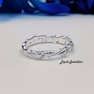 Puede incluir: Un anillo de plata con un diseño repetido de delfines. El anillo tiene un acabado pulido y el texto "Dipika Jewellers" es visible. El anillo se muestra sobre una superficie blanca.