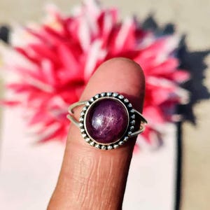 Puede incluir: Un anillo de plata con una gema grande, redonda y de color púrpura intenso. La gema está engastada en un bisel con un borde de cuentas. El anillo tiene un diseño de caña dividida. El fondo es rosa y blanco borroso.
