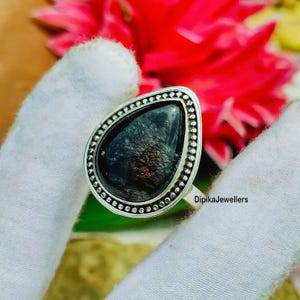 Op de afbeelding: Een zilveren ring met een druppelvormige, donkere edelsteen. De edelsteen heeft een iriserende glans met paarse en gouden tinten. De ring is versierd met kleine, cirkelvormige details. De tekst "DipikaJewellers" is zichtbaar.