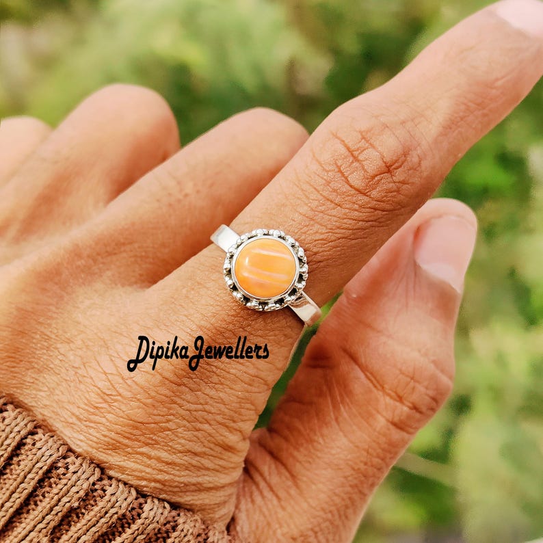 May include: A silver ring with a round, light orange gemstone. The gemstone is set in a decorative bezel. The ring is on a finger. The text "Dipika Jewellers" is visible.