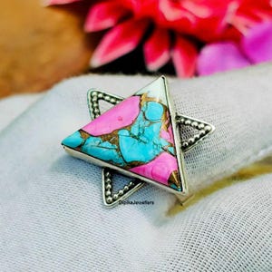 Op de afbeelding: Een zilveren ring met een driehoekige steen met een levendige mix van roze, turquoise en goud. De band van de ring heeft de vorm van een ster. De tekst "DipikaJewellers" is zichtbaar op de ring.