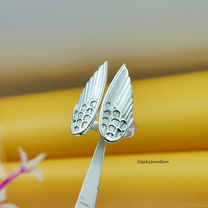 May include: A silver-colored ring with a wing design. The ring features two detailed wing shapes, each with textured feather patterns. The ring is held by a silver tool against a yellow background. The text "Dipikajewellers" is visible.