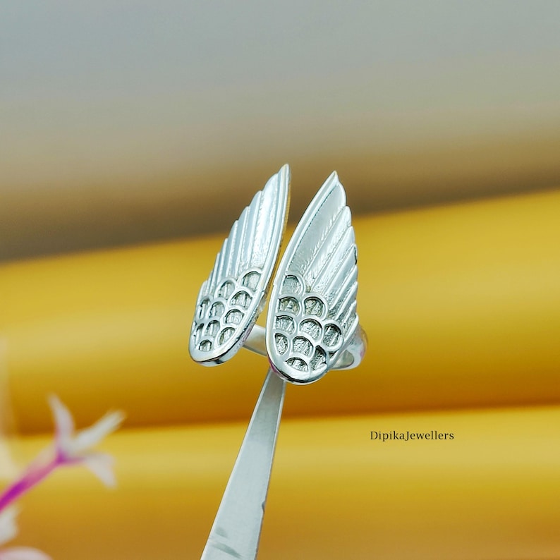 May include: A silver-colored ring with a wing design. The ring features two detailed wing shapes, each with textured feather patterns. The ring is held by a silver tool against a yellow background. The text "Dipikajewellers" is visible.