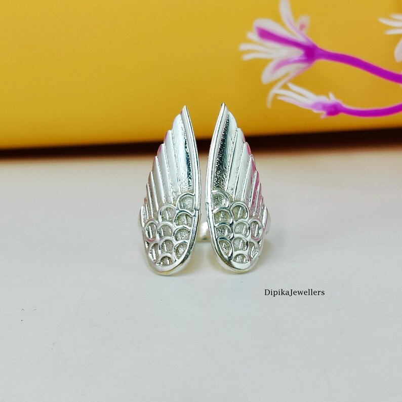 May include: A pair of silver-colored wing-shaped earrings. Each earring features a detailed feather design with a textured surface. The earrings are displayed on a light-colored surface with a yellow background and a purple flower.