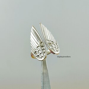 May include: A silver-colored ring with an open design featuring two stylized wing shapes. Each wing has detailed feather-like lines and a textured pattern. The ring is held by a pair of tweezers against a neutral background. The text "Dipikajewellers" is visible.