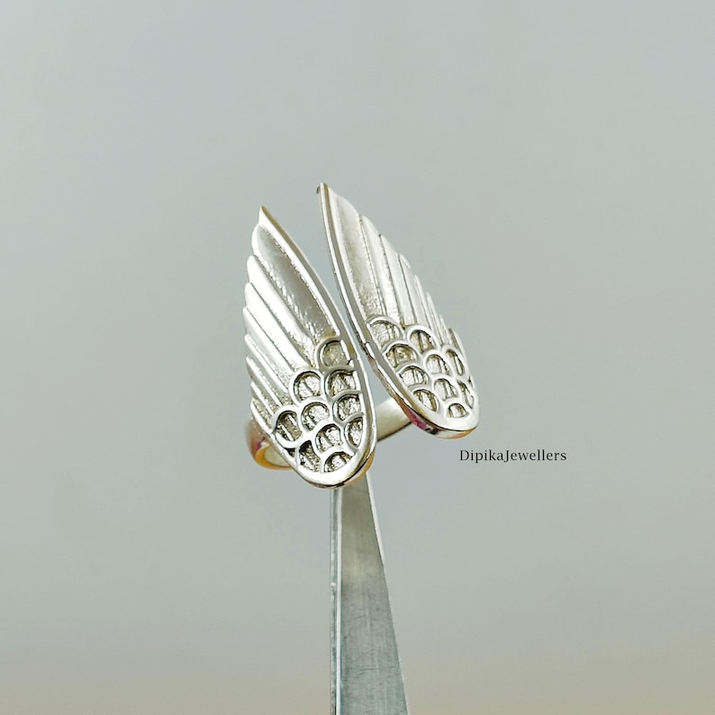 May include: A silver-colored ring with an open design featuring two stylized wing shapes. Each wing has detailed feather-like lines and a textured pattern. The ring is held by a pair of tweezers against a neutral background. The text "Dipikajewellers" is visible.