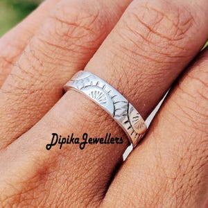 May include: A silver ring with a repeating sunburst pattern. The ring is worn on a finger. The text "Dipika Jewellers" is visible on the finger.