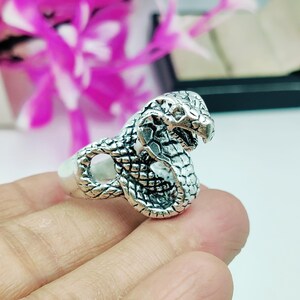 May include: A silver-colored ring in the shape of a cobra snake. The ring features detailed scales and a raised cobra head with an open mouth. The ring is shown on a finger against a blurred background of pink and white.