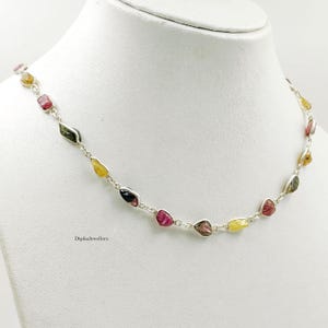 May include: A silver chain necklace featuring a series of small, irregularly shaped, raw tourmaline gemstones in various colors including pink, yellow, and green. The text DipikaJewellers is printed in small black letters on the lower left side.
