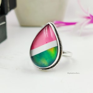 May include: A teardrop-shaped ring featuring a smooth, multi-colored inlay with distinct sections of pink, white, and a gradient of green and yellow. The stone is set in a polished silver frame with a twisted rope-style border and a plain band.