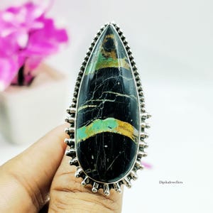 Puede incluir: Un anillo en forma de lágrima con una gran piedra negra pulida que presenta vetas horizontales de color turquesa y marrón dorado. La piedra está engastada en un bisel de plata esterlina decorado con un borde de cuentas y pequeñas garras.