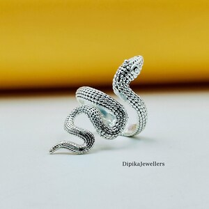 Könnte beinhalten: Ein silberner Schlangenring mit detaillierten Schuppen und einem erhobenen Kopf. Der Ring wickelt sich in einem spiralförmigen Design um den Finger, wobei sich der Kopf der Schlange oben befindet. Der Ring befindet sich auf einer weißen Oberfläche mit gelbem Hintergrund. Text: DipikaJewellers.