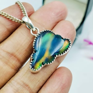 May include: A silver pendant necklace featuring a colorful, iridescent stone in shades of blue, green, and yellow. The stone is set in a silver frame with a decorative edge and hangs from a silver chain. The pendant is held in a hand.