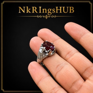 May include: A silver-toned ring featuring a large, oval-cut deep red gemstone set in a pronged mounting. The band displays an intricate, textured cross-hatched pattern. The text NkRingsHUB is displayed in gold lettering at the top of the image.