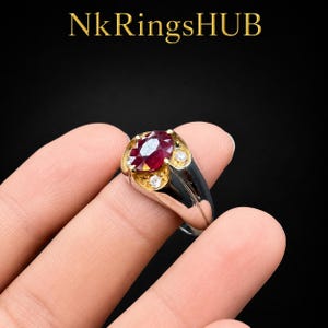 May include: A ring featuring a central oval-cut deep red gemstone set in a gold-toned bezel, flanked by two small clear round stones. The band is a polished silver-toned metal. The text NkRingsHUB is visible at the top of the image.