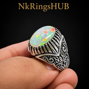 May include: A silver ring featuring a large, oval-shaped iridescent opal gemstone with flashes of orange and green. The band is crafted with intricate, dark-oxidized metal engravings and geometric patterns. The text NkRingsHUB is visible above.