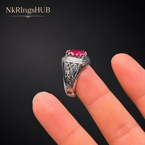 May include: A silver-toned ring featuring a faceted, oval-shaped deep red gemstone set in a prong setting. The band displays intricate, dark-toned scrollwork engravings along the sides. The text NKRingsHUB is visible in the upper left corner.