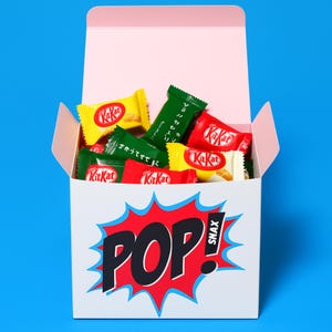 May include: A white box with a pink interior, filled with colorful Kit Kat candy bars. The box has a red and black "POP! SNAX" graphic on the front, with a blue background. The Kit Kat wrappers are yellow, green, and red.