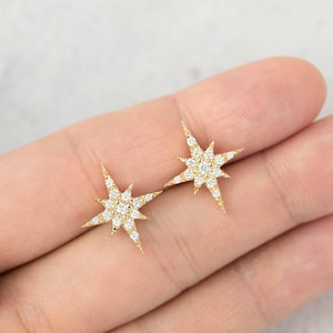 Puede incluir: Un par de pendientes de botón dorados con forma de estrella, cada uno con un grupo central de pequeñas piedras redondas y transparentes dispuestas en una estrella de múltiples puntas con rayos radiantes, creando un diseño celeste brillante.