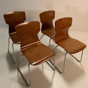 Könnte beinhalten: Vier Holzstühle mit geschwungenen Rückenlehnen und Sitzflächen, mit natürlicher Holzmaserung. Die Stühle haben ein freitragendes Metallgestell in Chromfarbe. Die Stühle sind in einer Gruppe angeordnet und zeigen ihr Design und ihre Konstruktion.