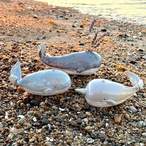 Pode incluir: Três figuras de baleias de cerâmica em tons de cinza e branco numa praia de seixos. Uma baleia tem lavanda. As baleias têm barbatanas detalhadas e um design minimalista.
