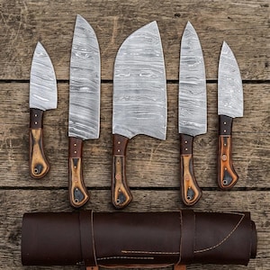 May include: A set of five kitchen knives with Damascus steel blades featuring a wavy pattern and brown wooden handles with decorative pins, arranged above a dark brown leather knife roll with stitched details and a strap closure.