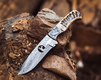 May include: A detailed hunting knife with a patterned blade and a handle made of bone-like material. The blade features a circular emblem with a deer silhouette. The knife rests on a rough, textured surface, suggesting an outdoor setting.