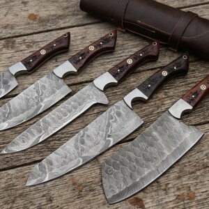 May include: A set of five kitchen knives with patterned Damascus steel blades and dark brown wooden handles featuring decorative metal rivets. The knives are arranged in a row with a dark brown leather roll case positioned at the top.