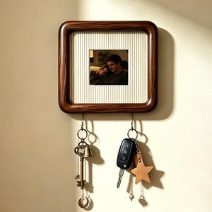 Op de afbeelding: Een vierkante houten sleutelhouder voor aan de muur met een donkerbruine lijst en een crèmekleurige achtergrond met krijtstrepen. In het midden zit een kleine foto van twee personen. Twee metalen haken hangen eronder met een oude sleutel en autosleutels.