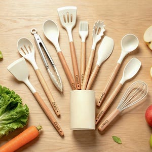 May include: A collection of cream-colored kitchen utensils with wooden handles, including spatulas, spoons, and a whisk, arranged on a wooden surface. The word "Grace" is printed on the handles. Fresh produce, such as apples, a lemon, and lettuce, are also present.