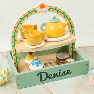 Può includere: Un set da tè giocattolo in legno con una teiera gialla, tazze e piattini su un vassoio a due livelli. Il vassoio include cupcake e una torta a forma di cuore. Il nome "Denise" è stampato. Motivi floreali e a foglie.