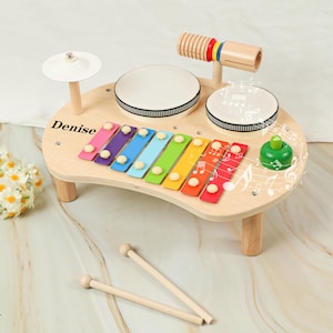 Può includere: Set di strumenti musicali giocattolo in legno con xilofono, tamburi e piatto. Lo xilofono ha tasti colorati e i tamburi sono bianchi con bordi neri. Il set include bacchette e il nome "Denise" è stampato sullo strumento.